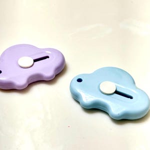 May include: Two cloud-shaped box cutters. One is light purple, the other is light blue. Each has a white circular slider and a small hole for a lanyard. The cutters are on a white surface.