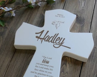 May include: White wooden cross with the name "Hadley" engraved in brown script. The cross includes a dove illustration and the text "Baptism July 16, 2017." A Bible verse is also inscribed on the cross.