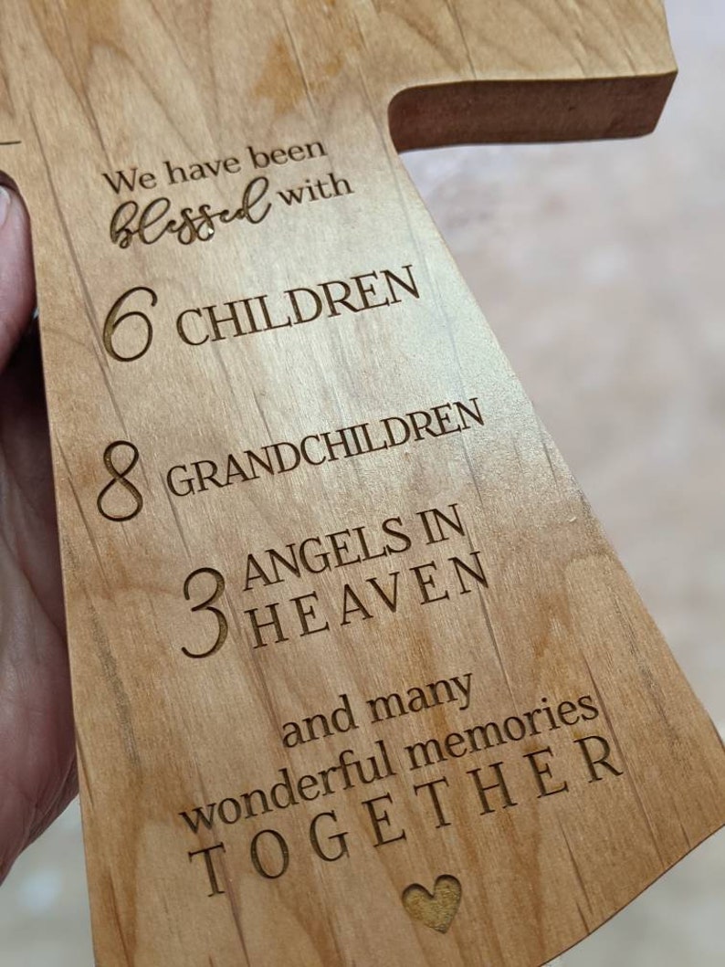 May include: Wooden cross-shaped plaque with engraved text. The text reads "We have been blessed with 6 CHILDREN, 8 GRANDCHILDREN, 3 ANGELS IN HEAVEN, and many wonderful memories TOGETHER" with a heart symbol at the bottom. The wood has a light brown color.