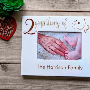 May include: White picture frame with the text "2 generations of love" and "The Harrison Family" in brown lettering. The frame has a photo of an older hand holding a baby's foot.
