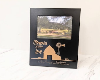 May include: Black wooden picture frame with a gold engraved inscription that reads "Memories are a way of holding on to the ones we love." The inscription also includes a barn, windmill, and tractor. The frame has a photo of a rural landscape with trees and a dirt road.