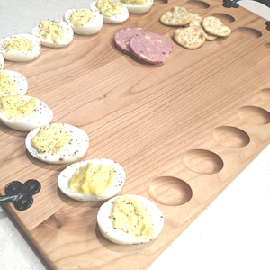 May include: A wooden serving board with a built-in egg holder and a handle. The board is filled with deviled eggs, crackers, and sliced meat.