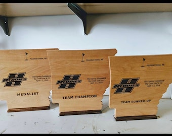 May include: Three wooden plaques shaped like the state of Arkansas, each with a different award inscription. The plaques are all brown and have a black and white logo that says "Reddies". The top plaque says "Medalist", the middle plaque says "Team Champion", and the bottom plaque says "Team Runner-Up".