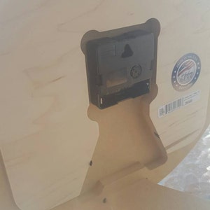 May include: The back of a wooden clock with a black clock mechanism and a sticker that says "Made in the USA" with a red, white, and blue American flag. The sticker also says "Rochester Dunn" and "Since 1990".