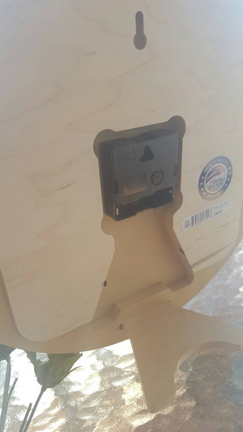 May include: The back of a wooden clock with a black clock mechanism and a sticker that says "Made in the USA" with a red, white, and blue American flag. The sticker also says "Rochester Dunn" and "Since 1990".