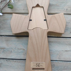 May include: A wooden cross-shaped photo frame with a natural wood finish. The back features a rectangular opening for a photo and a small label with text. The frame is placed on a weathered wooden surface.