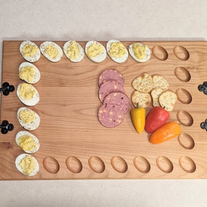 May include: A wooden serving board with black handles, featuring a variety of snacks, including deviled eggs, sliced meat, crackers, and peppers.