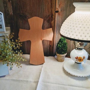 May include: A wooden cross with a light brown finish stands on a cream-colored tablecloth. The cross is the focal point, with a rustic wooden background. A vintage lamp with a white shade and floral accents is on the right.