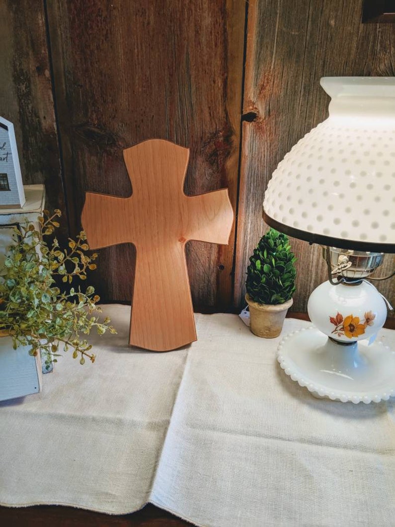 May include: A wooden cross with a light brown finish stands on a cream-colored tablecloth. The cross is the focal point, with a rustic wooden background. A vintage lamp with a white shade and floral accents is on the right.