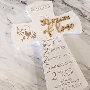 May include: A white wooden cross-shaped plaque with gold lettering and accents. The plaque commemorates a 50th wedding anniversary, with the names "Archie and Vickie Spangler" and the date "May 18, 1974-2024". It lists family members and pets.