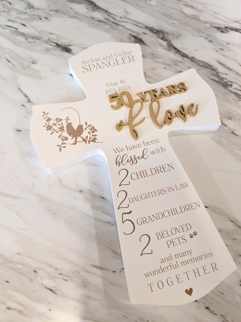 May include: A white wooden cross-shaped plaque with gold lettering and accents. The plaque commemorates a 50th wedding anniversary, with the names "Archie and Vickie Spangler" and the date "May 18, 1974-2024". It lists family members and pets.
