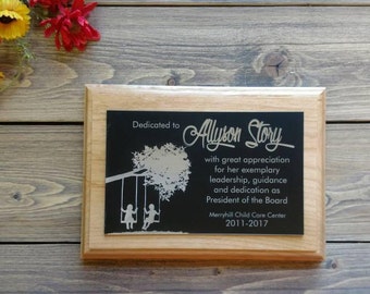 May include: A wooden plaque with a silver engraved inscription. The inscription reads "Dedicated to Alyson Story with great appreciation for her exemplary leadership, guidance and dedication as President of the Board Merryhill Child Care Center 2011-2017". The plaque features a silver engraved image of a tree with two children on swings.