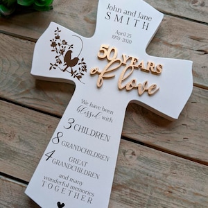 May include: White wooden cross-shaped plaque commemorating a 50th wedding anniversary. The plaque features the names "John and Jane SMITH" and the date "April 25 1970-2020". It includes the words "50 YEARS of love" and lists family members.