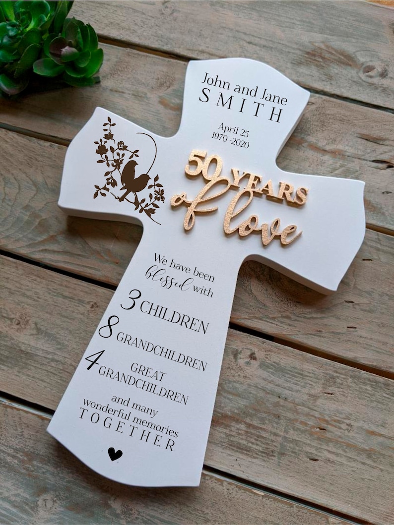 May include: White wooden cross-shaped plaque commemorating a 50th wedding anniversary. The plaque features the names "John and Jane SMITH" and the date "April 25 1970-2020". It includes the words "50 YEARS of love" and lists family members.