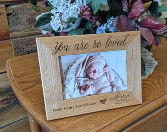 May include: A wooden picture frame with the text "You are so loved" engraved on the top. The frame holds a photo of a baby sleeping with a stuffed animal. The text "Noah Henry Lee Johnson" and "Blessed With You On September 11, 2023" is engraved below the photo.