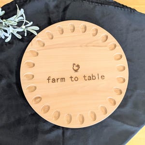 May include: A round, light-colored wooden egg holder with 20 oval-shaped indentations. The words "farm to table" are engraved on the surface, along with a rooster illustration. The holder sits on a black surface, with a sprig of greenery in the upper left corner.
