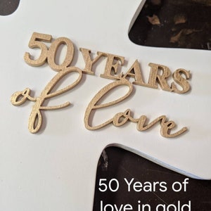 May include: A white wooden cross with gold-colored wooden letters that read "50 YEARS of Love". The text "50 Years of love in gold" is printed in black at the bottom of the cross.
