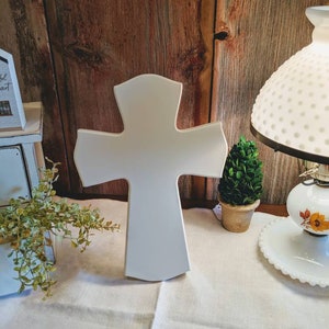 May include: A white wooden cross with a rounded top and flared arms stands on a cream-colored surface. The cross is the focal point, set against a rustic wooden background. A small potted plant and a vintage lamp are visible.
