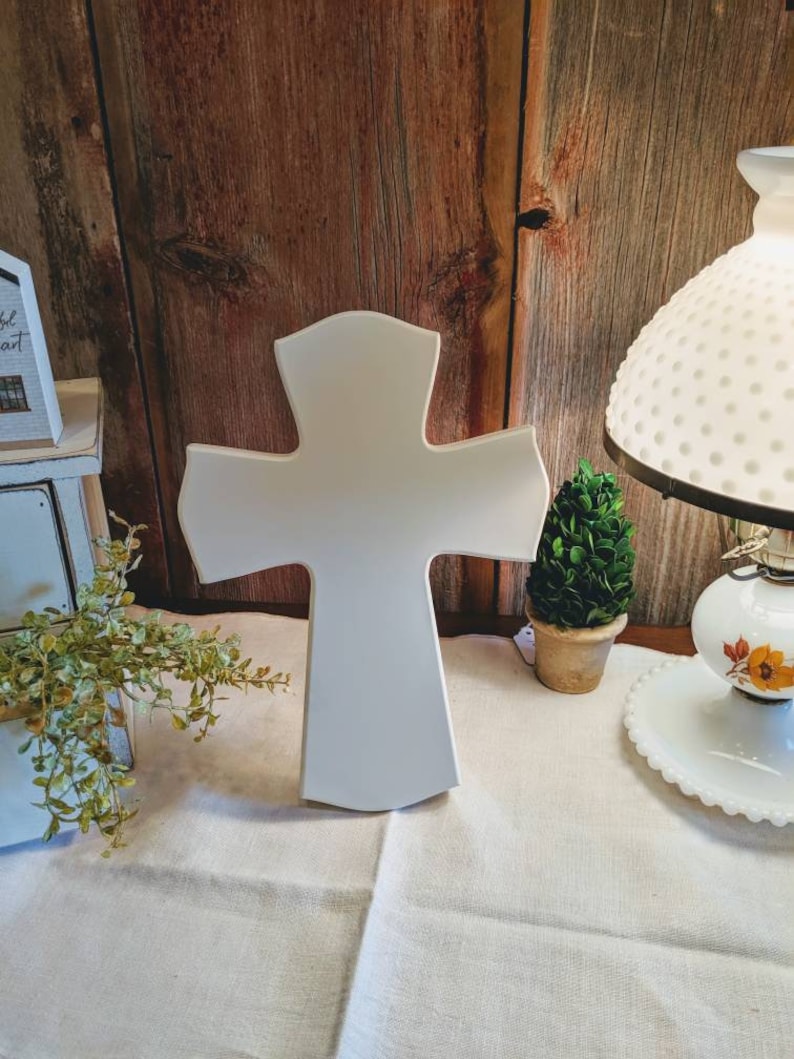 May include: A white wooden cross with a rounded top and flared arms stands on a cream-colored surface. The cross is the focal point, set against a rustic wooden background. A small potted plant and a vintage lamp are visible.