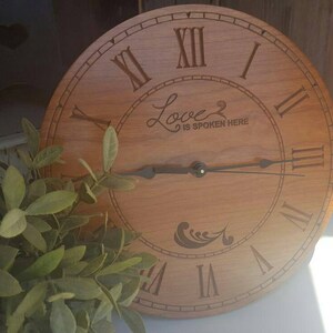 May include: A wooden clock with Roman numeral markings and a cursive inscription that reads "Love is spoken here".