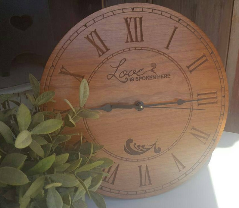 May include: A wooden clock with Roman numeral markings and a cursive inscription that reads "Love is spoken here".