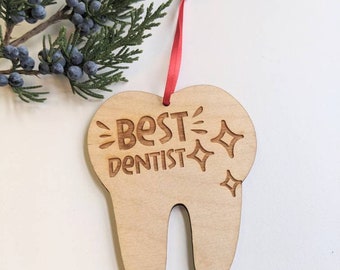 May include: A wooden tooth-shaped ornament with the text "Best Dentist" and sparkling stars. The ornament is attached to a red ribbon.