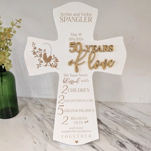 May include: A white wooden cross-shaped sign with the text "Archie and Vickie Spangler" and "50 Years of Love." It includes the dates 1974-2024 and the words "blessed with 2 children, 2 daughters-in-law, 5 grandchildren, 2 beloved pets." A decorative floral design is on the left.