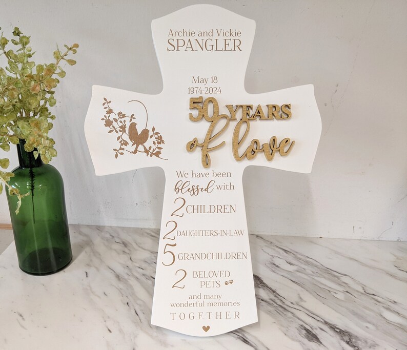 May include: A white wooden cross-shaped sign with the text "Archie and Vickie Spangler" and "50 Years of Love." It includes the dates 1974-2024 and the words "blessed with 2 children, 2 daughters-in-law, 5 grandchildren, 2 beloved pets." A decorative floral design is on the left.