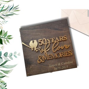 May include: A wooden keepsake box with gold lettering and a bird design, celebrating 50 years of love and memories. Includes the names James & Caroline and the years 1975-2025. Two cream-colored envelopes are also visible.