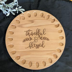 May include: A round wooden serving tray with a natural wood finish. The tray has 24 small indentations around the edge. The tray has the text "thankful & blessed" engraved in the center.