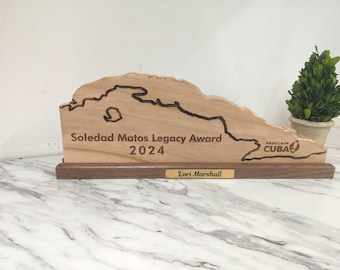 May include: A wooden award plaque in the shape of Cuba with the text "Soledad Matos Legacy Award 2024" and "Lori Marshall" engraved on it. The plaque is on a wooden base with a small potted plant to the right.