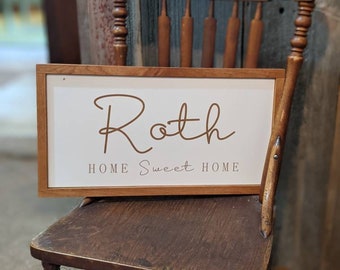 May include: A white wooden sign with a brown wooden frame. The sign says "Roth" in cursive script and "Home Sweet Home" in a smaller font. The sign is sitting on a wooden chair.