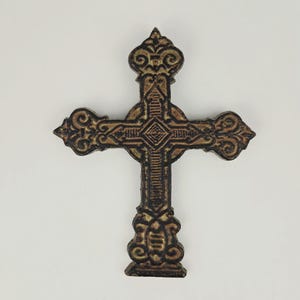 May include: Ornate, antique-style cross in shades of brown and gold. The cross features detailed scrollwork and a diamond-shaped center design. The religious symbol is made of a dark, possibly metal material, and is set against a plain white background.