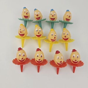 May include: A collection of vintage clown-themed food picks. Each pick features a clown face with a red smile, painted eyes, and a small hat. The picks have different colored bases: green, yellow, and red.