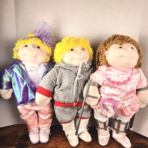 May include: Three vintage Cabbage Patch Kids dolls. The dolls have yellow or brown yarn hair and are wearing various outfits, including a pink satin tracksuit, a gray sweatsuit, and a pink dress. One doll has a crutch and a purse.