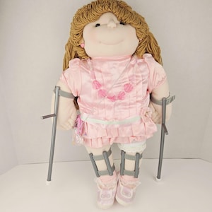 May include: A plush doll with light brown hair, wearing a pink dress, pink shoes, and a pink beaded necklace. The doll has braces on its legs and is using two silver crutches for support. The doll has a smiling expression.