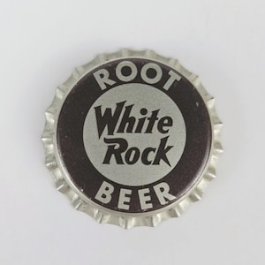 May include: A silver bottle cap with a black background. The text "ROOT BEER" is printed in white letters around a circle. Inside the circle, the text "White Rock" is printed in black letters.