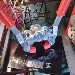 May include: A silver metal bottle capper with red handles and jaws, resting on a black digital scale. The scale displays a weight of 1.084. A ruler is visible to the right. The capper is designed for sealing bottles.