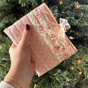May include: A pink and white floral fabric item with a bow. The item features a central panel with pink roses and green leaves on a cream background. The item is held in a hand, with a Christmas tree in the background.