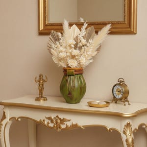 May include: A decorative arrangement featuring a green vase with a brown rim and a leather strap, filled with dried flowers. A small gold statue, a gold clock, and a gold tray are on a white table with gold trim. A gold framed mirror is in the background.