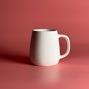 Pode incluir: Uma caneca de cer&acirc;mica branca com corpo arredondado e al&ccedil;a. A caneca est&aacute; sobre um fundo vermelho s&oacute;lido.