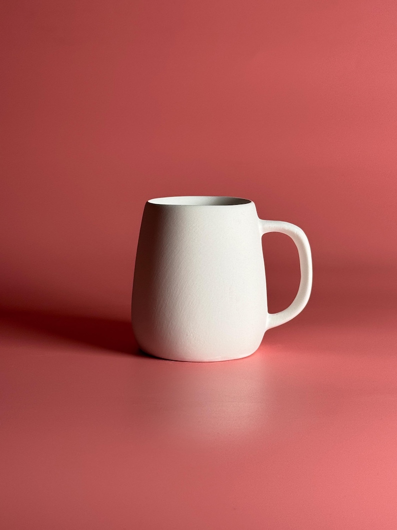 Pode incluir: Uma caneca de cer&acirc;mica branca com corpo arredondado e al&ccedil;a. A caneca est&aacute; sobre um fundo vermelho s&oacute;lido.