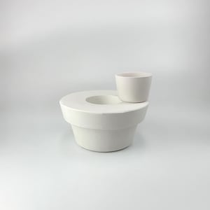 Puede incluir: Maceta de cerámica blanca con una taza de cerámica blanca más pequeña encima. La maceta tiene una abertura circular en el centro.