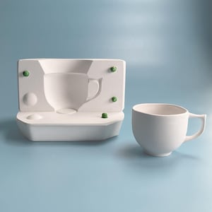 Könnte beinhalten: Eine weiße Keramik-Teetasse mit Henkel steht neben einer weißen Form mit einer tasseförmigen Vertiefung. Die Form hat grüne Akzente. Der Hintergrund ist einfarbig hellblau.