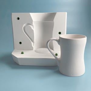 Puede incluir: Una taza de cerámica blanca con asa, junto a un molde de yeso blanco de una taza con asa. El molde tiene varios círculos verdes pequeños en los lados.