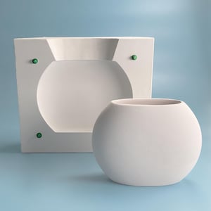 May include: A white ceramic mold for making a round vase. The mold has three green dots on the front and a round opening in the center. A white ceramic vase is sitting next to the mold.