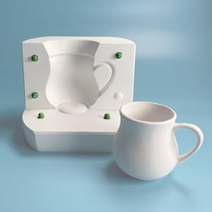 May include: A white ceramic mug with a rounded body and a handle. The mug is next to a white plaster mould of a similar mug.
