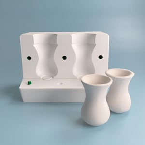 Może przedstawiać: Biała ceramiczna forma z trzema odciskami wazonów, obok dwóch gotowych białych wazonów. Forma ma zielone akcenty, a wazony mają klasyczny, elegancki kształt. Tło jest jednolicie jasnoniebieskie.