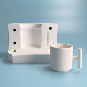 May include: A white ceramic mug with a unique T-shaped handle sits next to a white plaster mold. The mold has a rounded opening and several small holes.