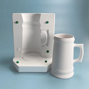 May include: A white ceramic beer stein with a handle and a mold for making the stein. The mold is made of white plaster and has several green dots on it.